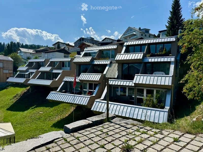 Modern multi-story building with a unique, tiered design featuring large windows and balconies, surrounded by a well-maintained lawn and trees.