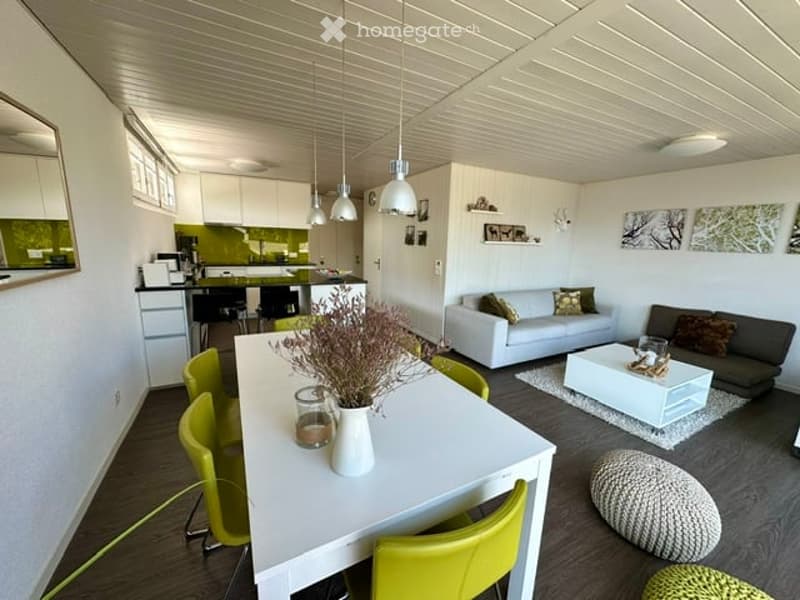 Open-plan living and dining area with modern furniture, including a white sofa, grey armchair, white coffee table, white dining table with green chairs, and a small kitchen area with white cabinets and a green backsplash.