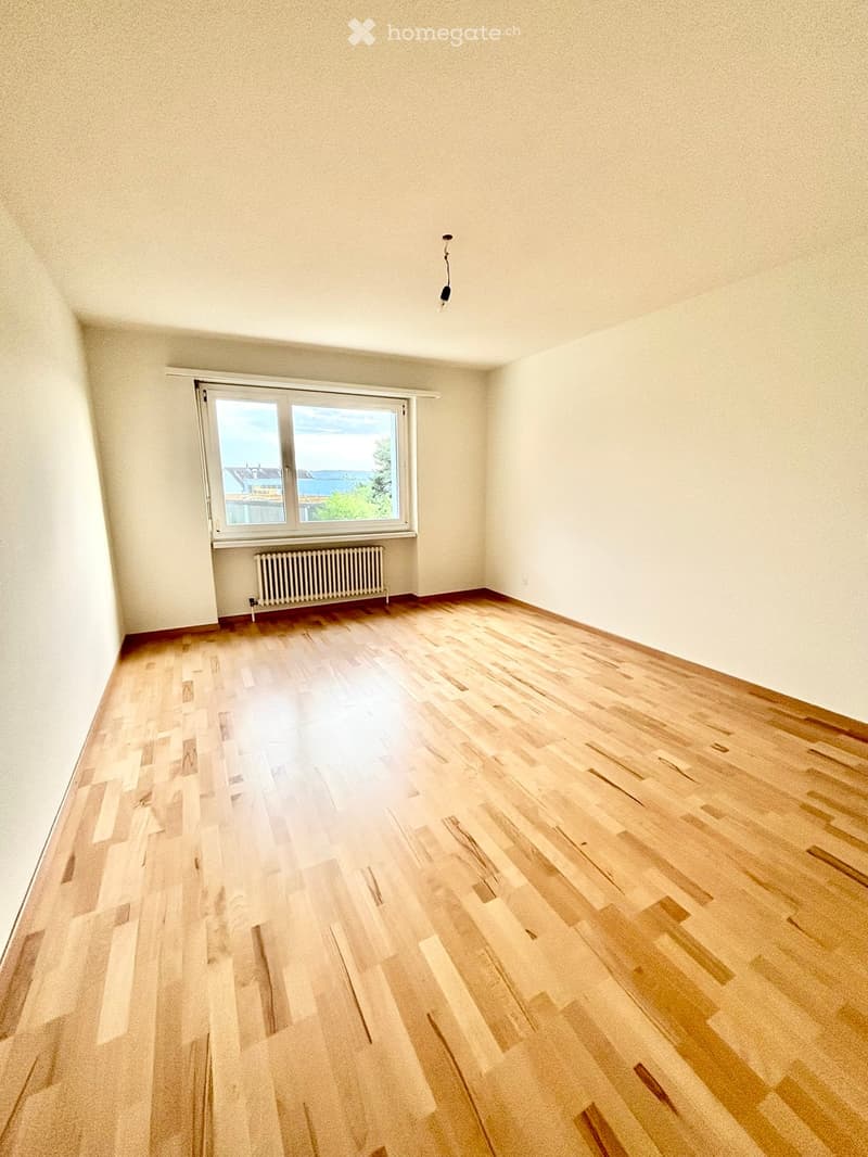 Empty room with wooden flooring, white walls, and a large window offering a view of the sea and surrounding landscape.