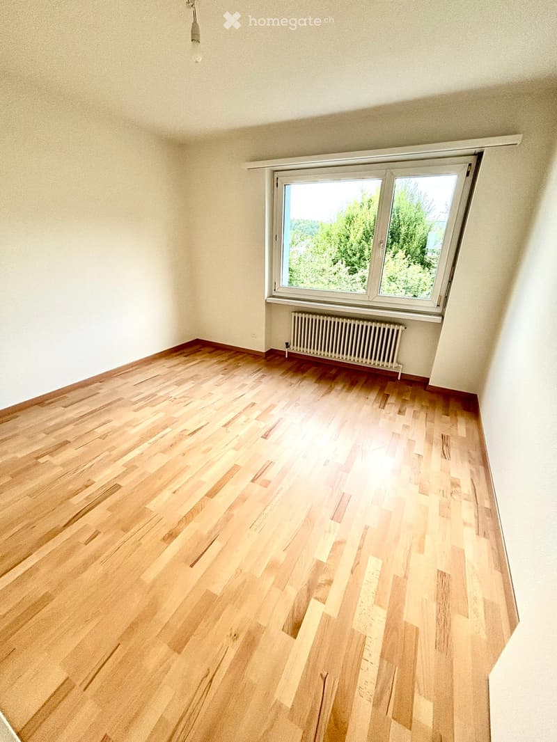 Empty room with wooden flooring, white walls, and a large window with a view of greenery. A radiator is visible beneath the window.