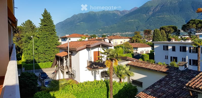Houses with tiled roofs, mountains in the background, trees, bushes, car parked in front