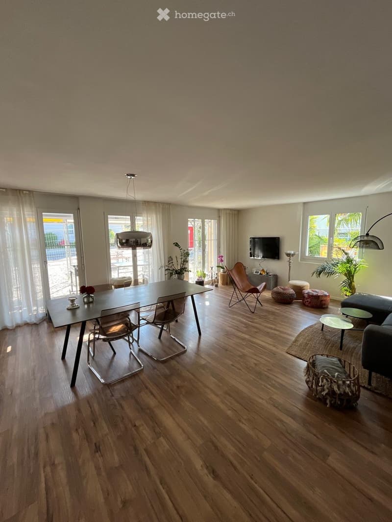 Open-plan living and dining area with wooden flooring, large windows, and modern furniture including a sofa, dining table, chairs, and decorative plants.