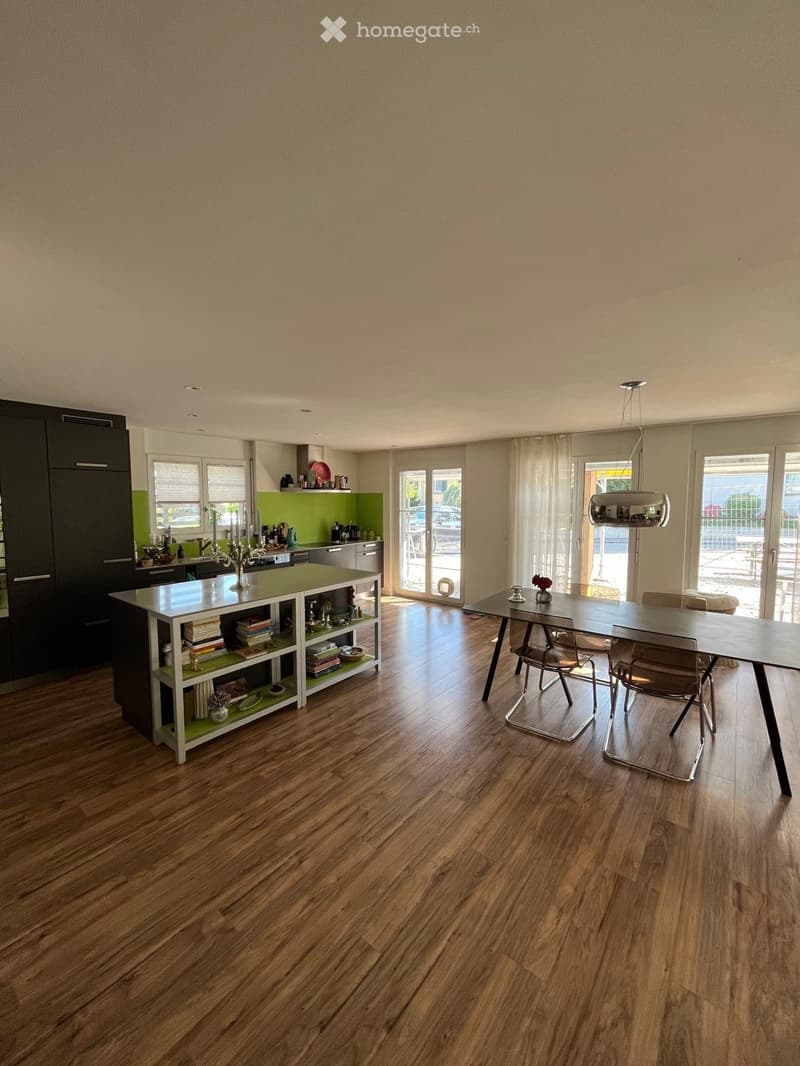 Open-plan kitchen and dining area with wooden flooring, modern kitchen island, dining table, and chairs. Features include large windows with curtains, a hanging pendant light, and various kitchen items on the island and shelves.