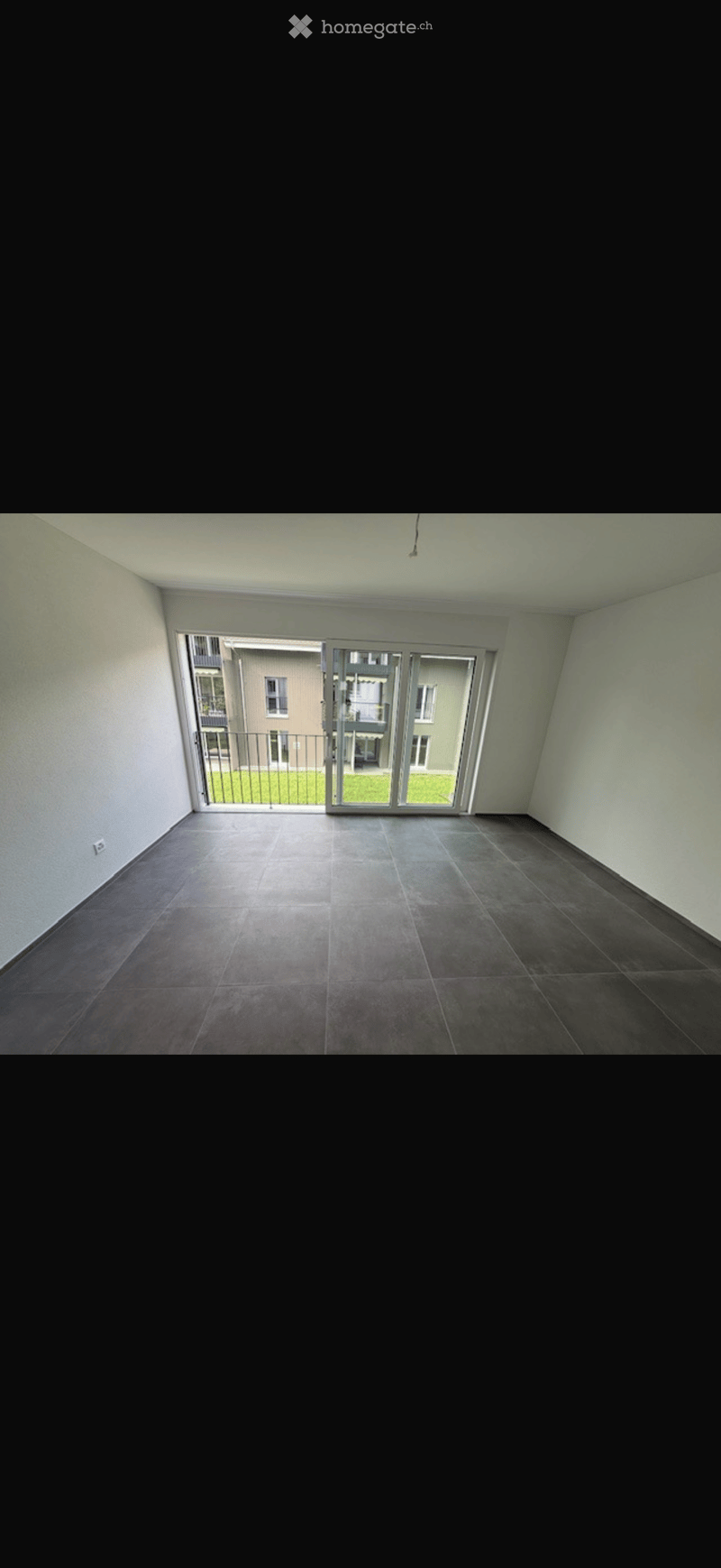 Empty room with dark tiled floor, white walls, and large sliding glass doors leading to a balcony with a view of a building and green area.