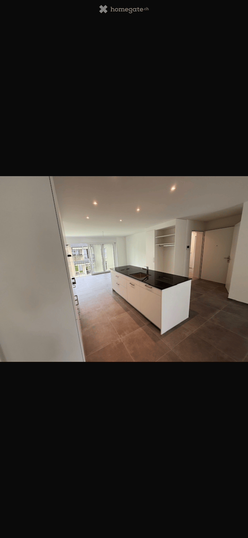 Empty kitchen with white cabinets, black countertop, and tiled floor. Large windows and a door leading to a balcony or outdoor area are visible.