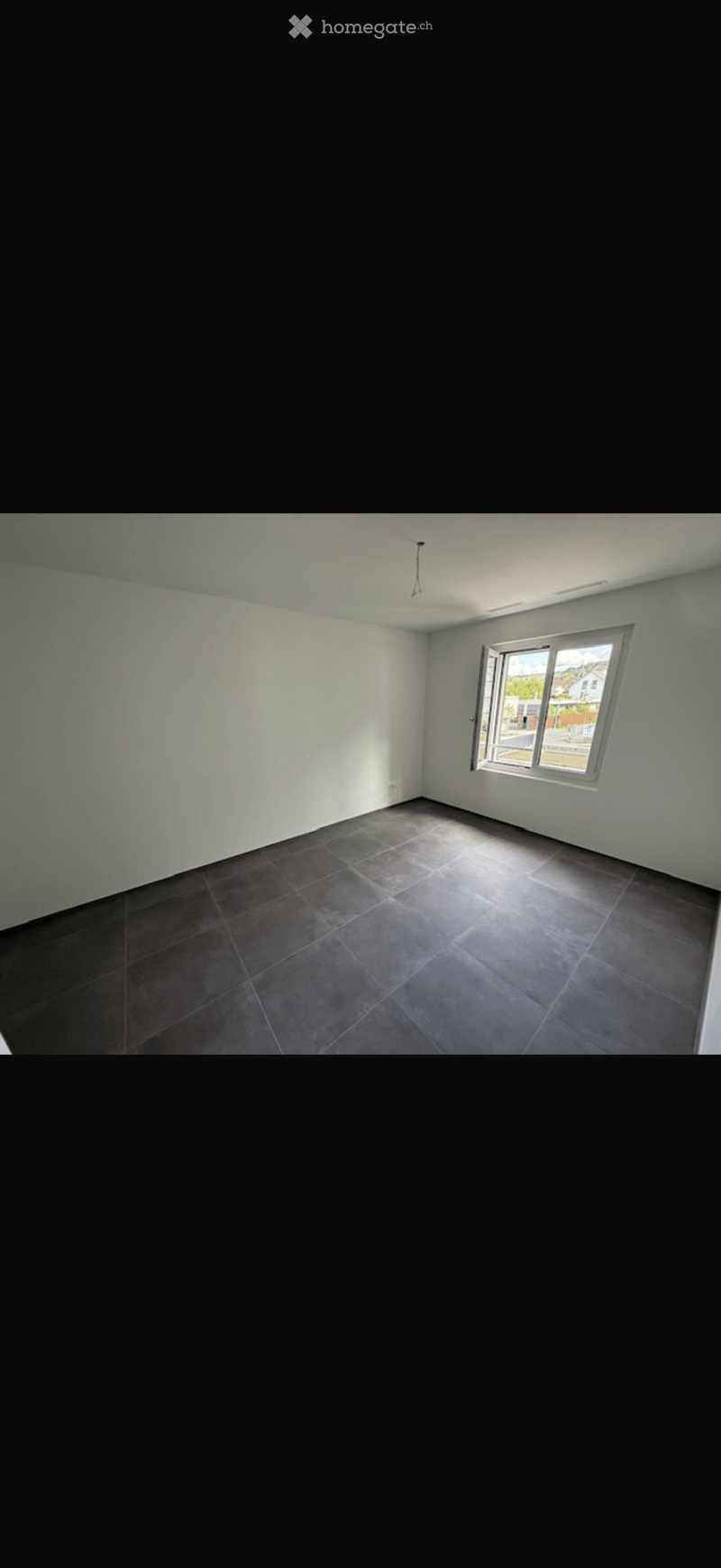 Empty room with light-colored walls, dark tiled floor, and a large window letting in natural light.