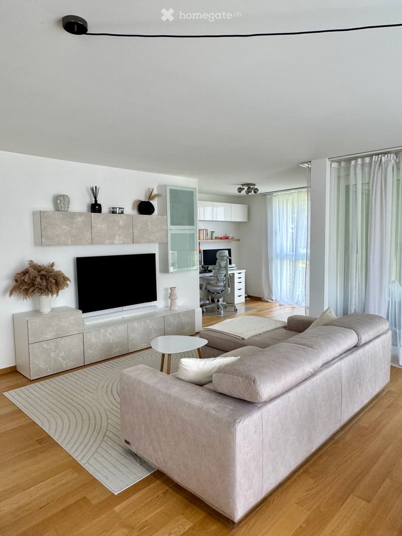 Modern living room with a grey sectional sofa, white coffee table, TV stand, and a small office area with a chair and desk.