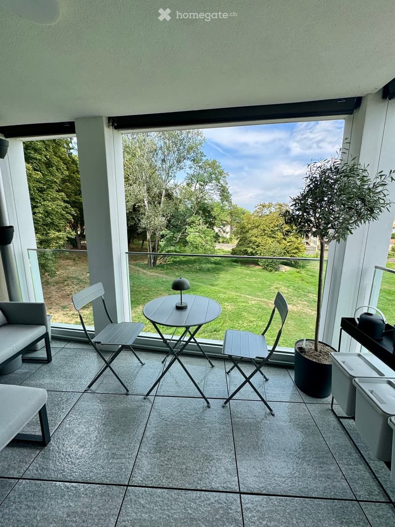 Modern balcony with grey tiled floor, glass railing, grey folding chairs, small round table with lamp, grey sofa, potted tree, and scenic view of green lawn and trees.