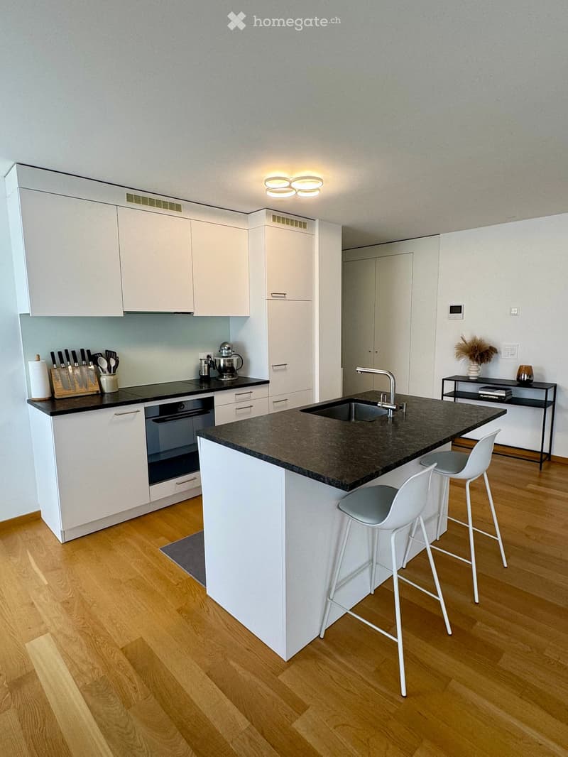 Modern kitchen with white cabinets, dark granite countertop, island, sink, and two white stools. Includes a knife block, blender, and a small table with a vase.