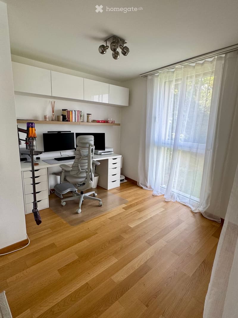 Modern, well-lit home office with wooden flooring, large window with white curtains, ergonomic chair, desk with computer and monitor, built-in white cabinets, and decorative items on shelves.