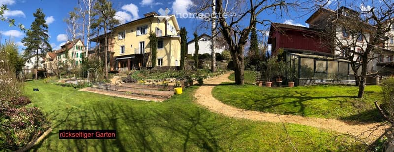 A well-maintained garden with a lawn, pathways, potted plants, and several residential buildings in the background.