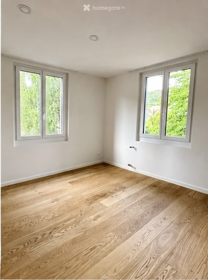 Completely empty room with bare white walls and wooden floor. Two windows with white frames and blinds.