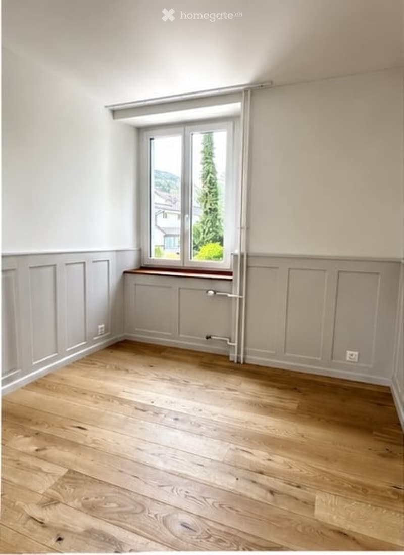 Empty room with wooden flooring, white walls, and a large window with a view of greenery outside.