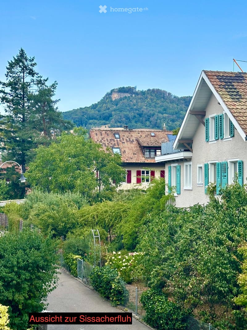 Two-story house with red-tiled roof, green shutters, and white walls, surrounded by lush greenery and trees, with a scenic mountain view in the background.