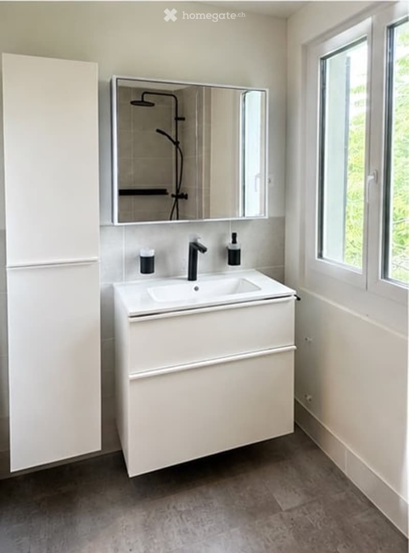 Modern bathroom with a white sink, mirror, and cabinet. The sink has a sleek black faucet and a small bottle on the counter. The walls are white, and the floor is a dark gray tile.