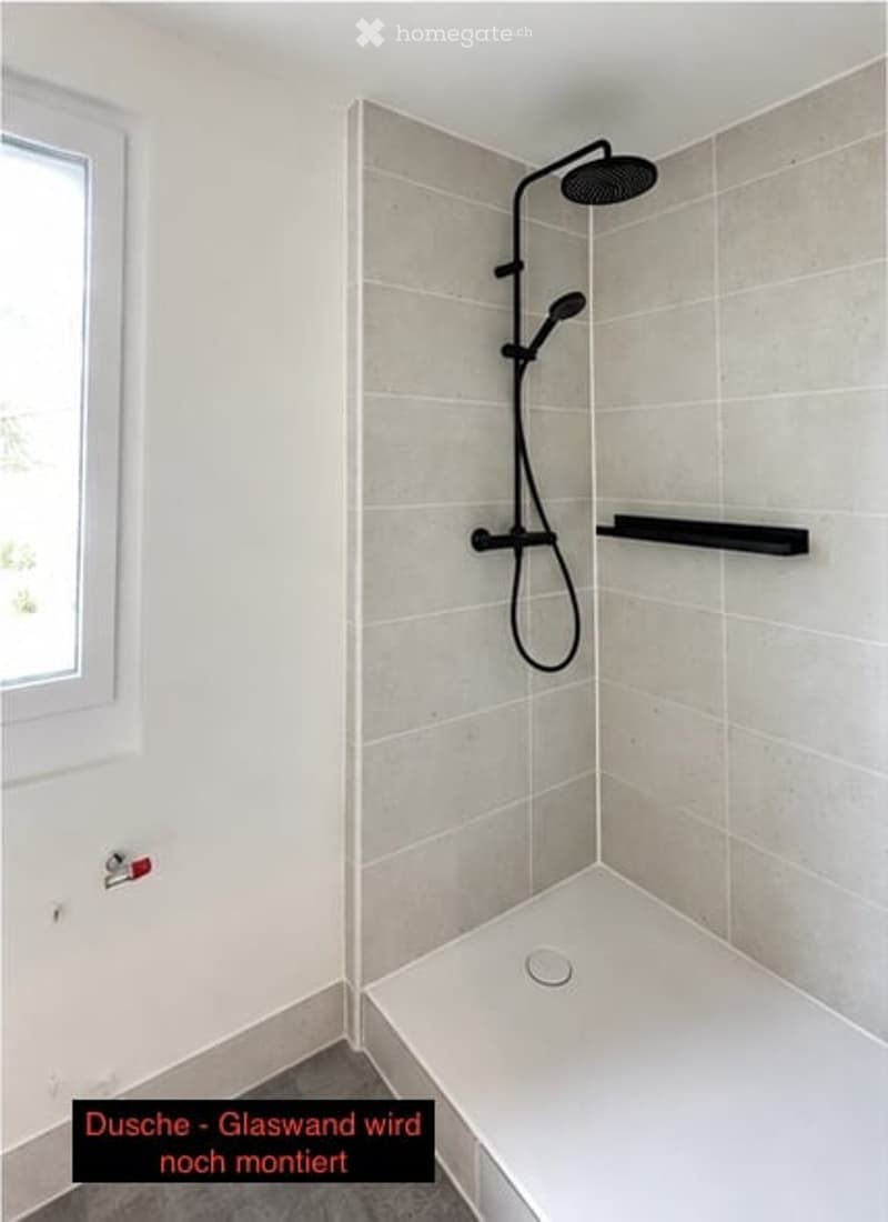 Modern shower area with light grey tiles, a showerhead, a hose, and a black shelf. The glass wall is not yet installed.