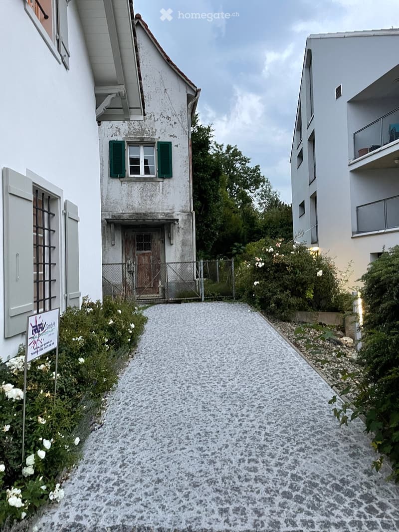 A narrow cobblestone pathway leads to a small house with a wooden door and green shutters, flanked by modern buildings and greenery.