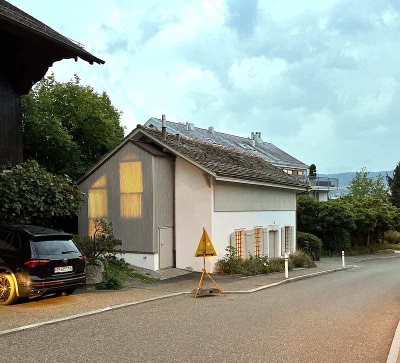 Two-story house with a modern design, featuring a garage, a driveway, and a parked car. The house has a combination of light-colored walls and dark roofing, with visible windows and a small garden area.