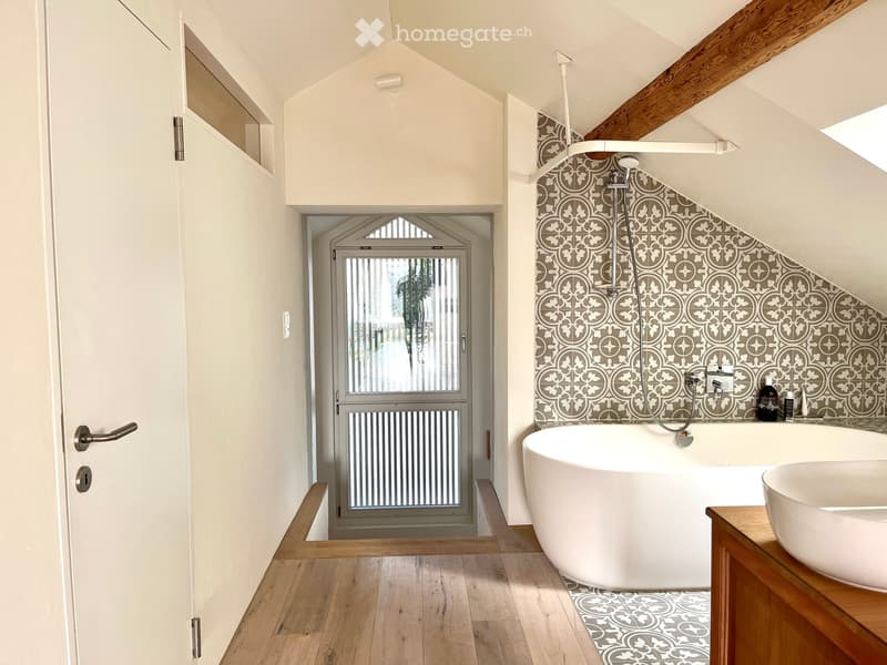 Modern bathroom with a freestanding bathtub, sink, and showerhead. The walls feature decorative tiles, and the floor is wooden.