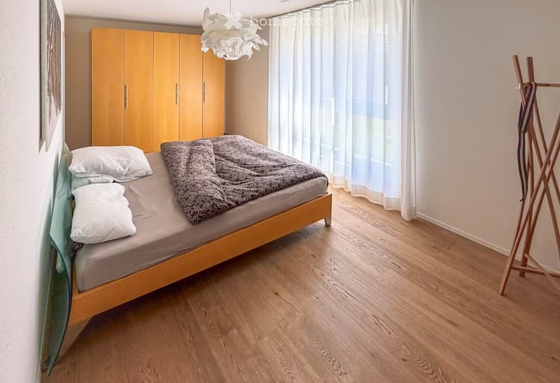 A modern bedroom with a wooden bed, pillows, a wardrobe, and a coat rack. The room features light wooden flooring and large windows with white curtains.