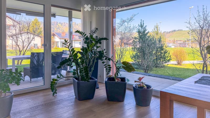 Bright living room with large windows, wooden floor, and several potted plants. A small table and chairs are visible near the window, and the view shows a green landscape with trees and houses.