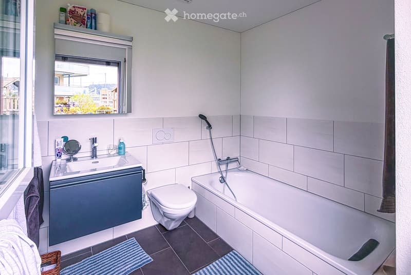 Modern bathroom with white tiles, a bathtub, toilet, sink, and a mirror above the sink. The room features a window with a view, a towel on the floor, and a small shelf with toiletries.