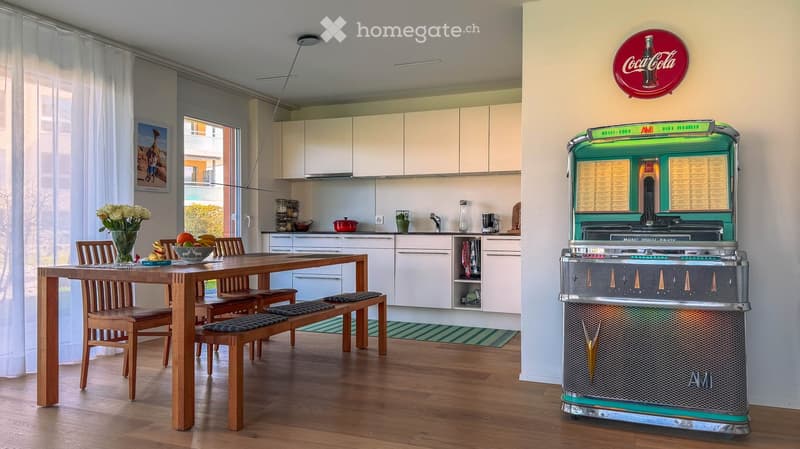 A modern kitchen and dining area with a wooden dining table, chairs, a vase with flowers, a retro-style refrigerator, and a sink area with various items.