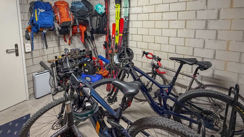 Storage area with multiple bicycles, backpacks, skis, and helmets neatly arranged against a brick wall.