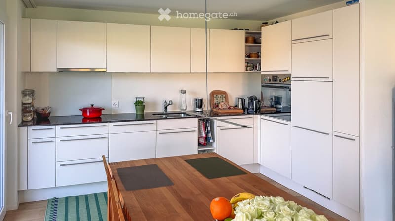 Modern kitchen with white cabinets, stainless steel appliances, and a wooden dining table with chairs. The counter features a sink, a red pot, a blender, and a coffee maker. A fruit arrangement with oranges, bananas, and roses is on the table.