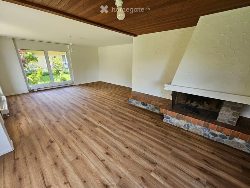 Empty room with wooden flooring, a fireplace with a stone surround, and large windows offering a view of the garden.