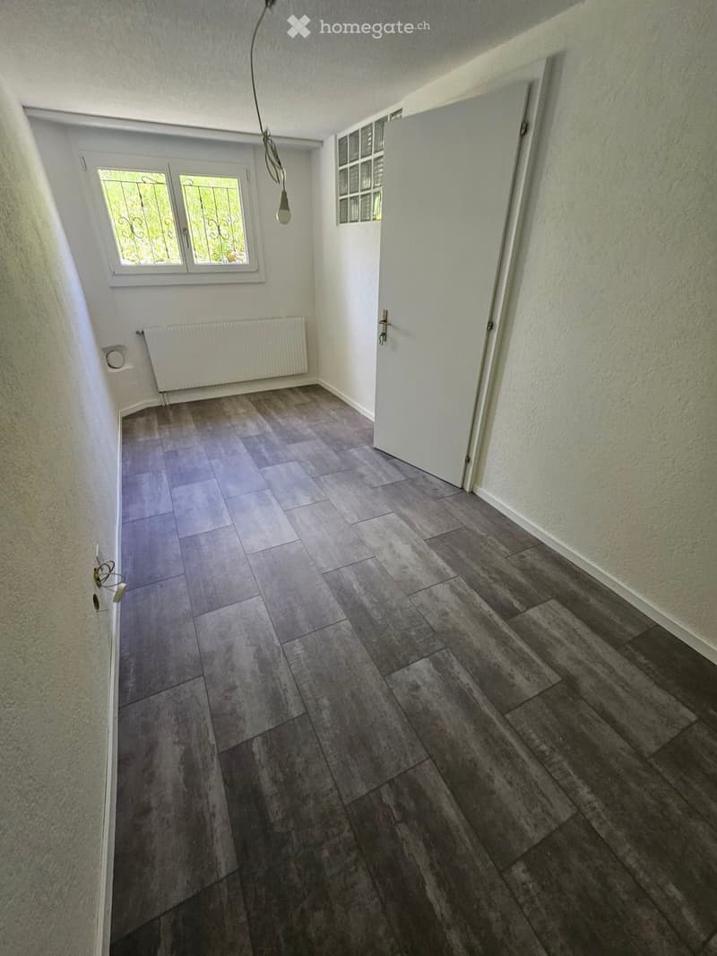 Empty hallway with tiled and wooden flooring, white walls, a window with decorative bars, a radiator, and a pendant light fixture.