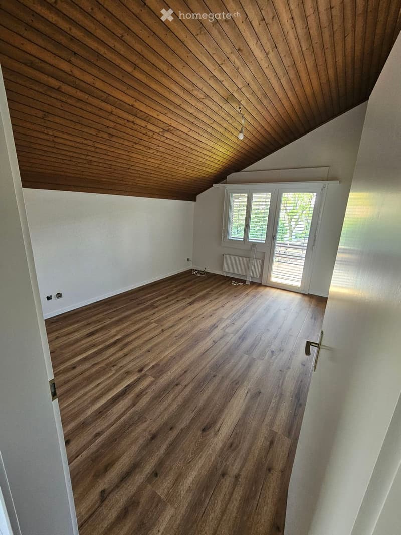 Empty room with wooden flooring, white walls, and a slanted wooden ceiling. Features a large window with blinds and a radiator under it.