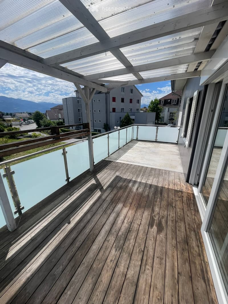 Wooden deck with glass railing, covered by a white pergola, overlooking a residential area with mountains in the background.