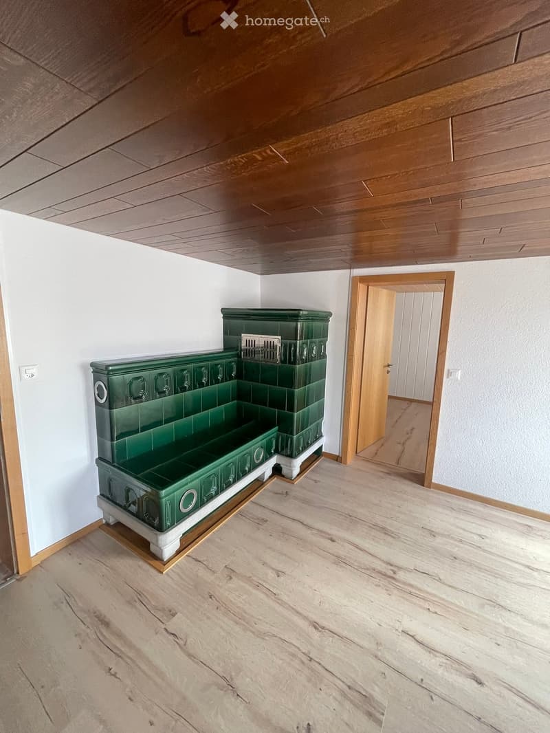 Empty room with wooden flooring, white walls, and a green fireplace in the corner. There is a doorway leading to another room.