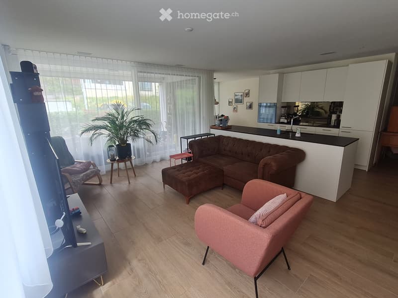 Spacious open-plan living room with large windows, hardwood flooring, brown tufted sofa, pink armchair, coffee table, and a kitchen area with white cabinets and countertops.