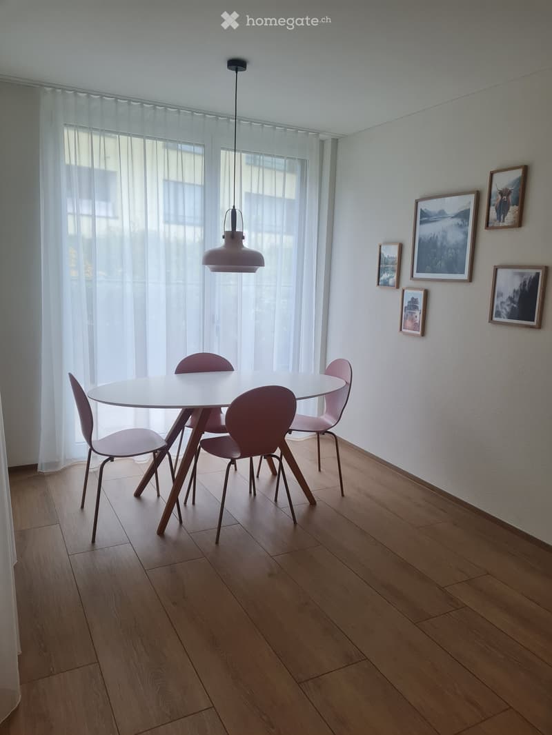 Spacious dining area with a round white table and burgundy chairs. Large windows provide natural light and a view of the balcony. The walls are decorated with framed artwork and photographs.