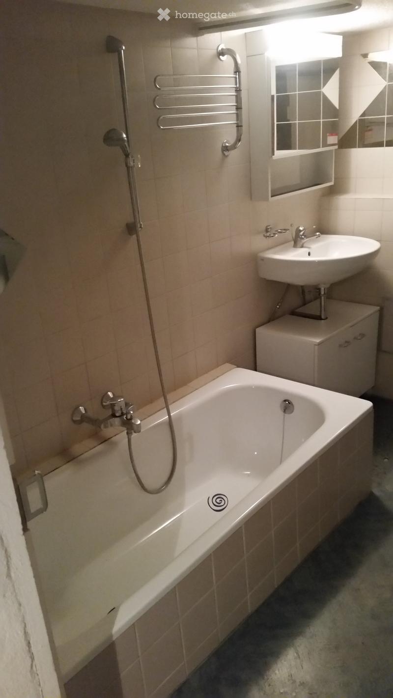 Modern bathroom with a bathtub, sink, mirror, and shower head. The walls are tiled, and there is a towel rack near the shower area.