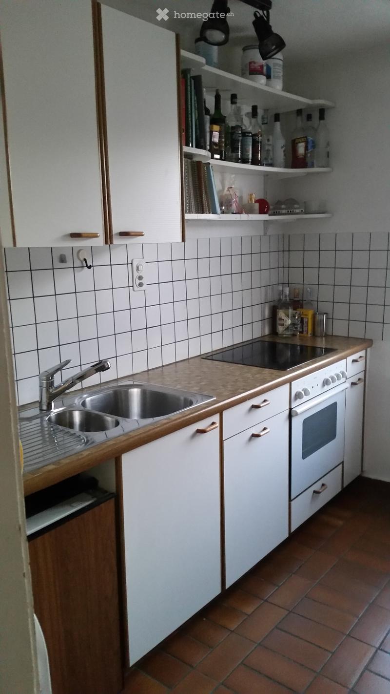 A compact kitchen with white cabinets, a tiled backsplash, a stainless steel sink, and an electric stove. Shelves hold various bottles and items.