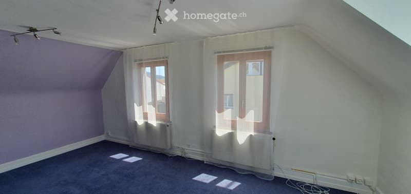 Empty room with slanted ceiling, carpeted floor, white walls, and large windows with sheer curtains. No furniture or equipment present.
