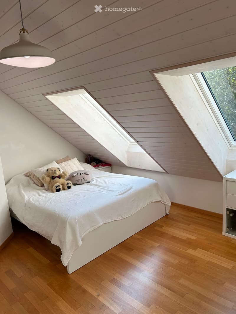 Cozy attic bedroom with wooden floors, slanted wooden ceiling, skylights, and a bed with pillows and stuffed animals.