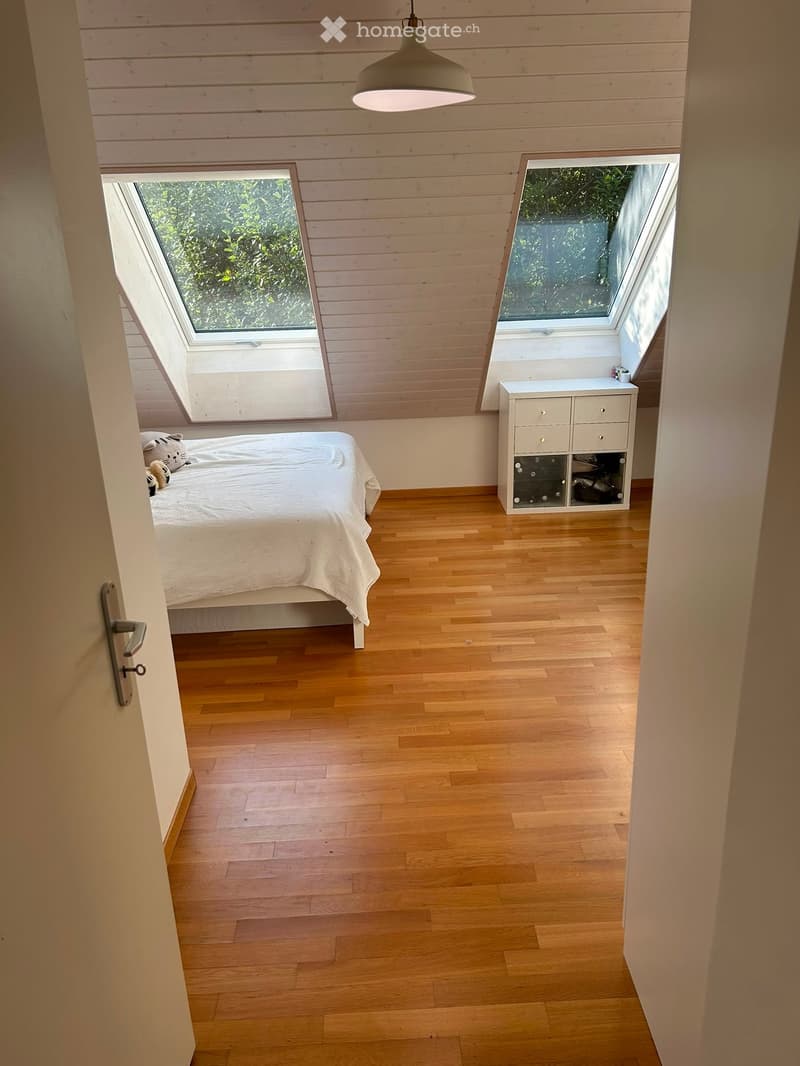 Bright, airy bedroom with wooden floors, slanted wooden ceiling, large skylights, a neatly made bed, and a small white storage unit.