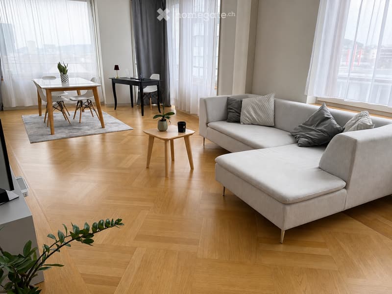 Modern living room with light wooden flooring, large windows with sheer curtains, grey sectional sofa, coffee table, side table, dining table with chairs, and a small desk with a laptop.