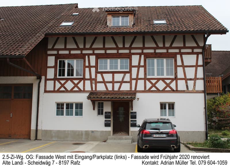 Two-story traditional house with white walls, wooden beams, multiple windows, and a tiled roof. Features a wooden front door, a small balcony on the upper floor, and a parked car in front.