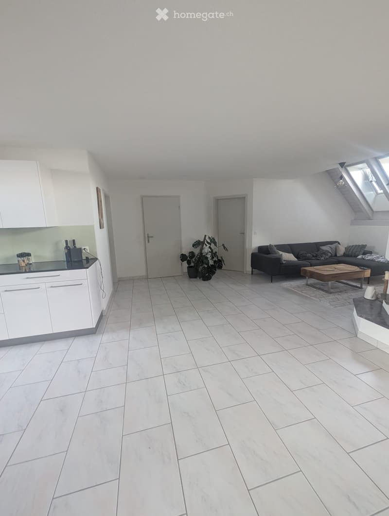 Open-plan living and kitchen area with white tiled flooring, white kitchen cabinets, a dark grey sofa, a wooden coffee table, and a potted plant.