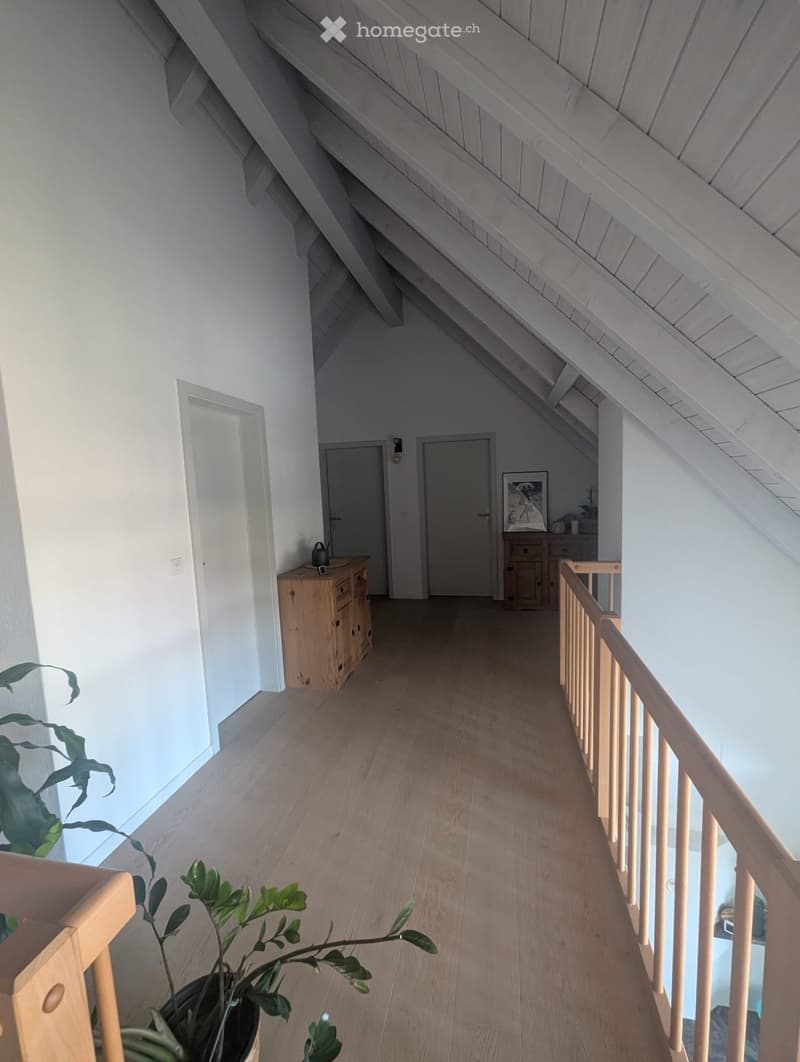 Bright, airy hallway with wooden flooring, white walls, and a slanted wooden ceiling. Features a wooden railing on one side and a small wooden cabinet with drawers on the left.