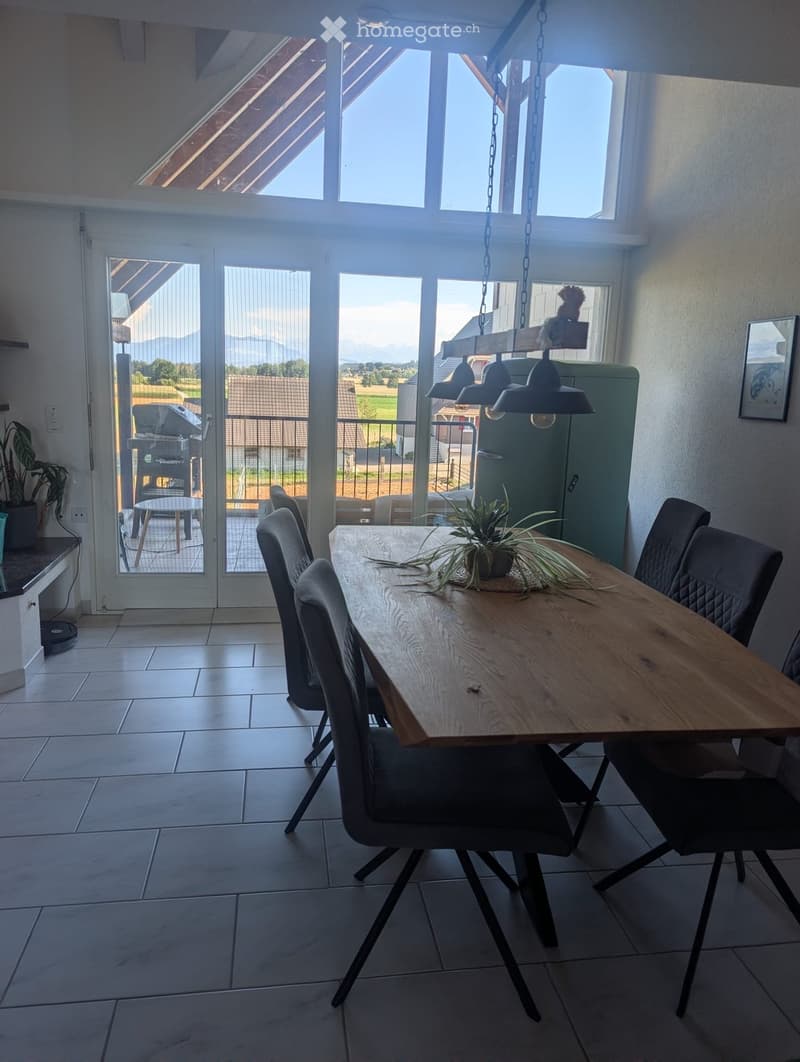 Bright dining area with a wooden table, six chairs, pendant lights, and a potted plant. Large windows and sliding doors lead to an outdoor patio with a grill and scenic view.