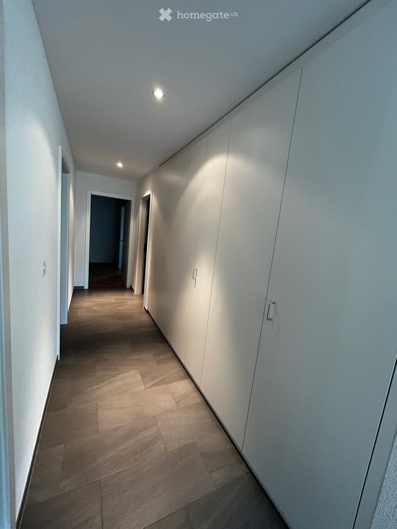 Empty hallway with light-colored walls, wooden flooring, and recessed lighting. Doors are visible at the end of the hallway.