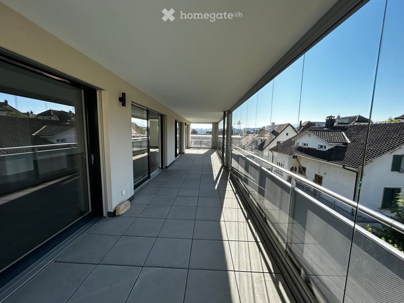 Spacious balcony with tiled floor, glass railings, and a view of neighboring rooftops and buildings.
