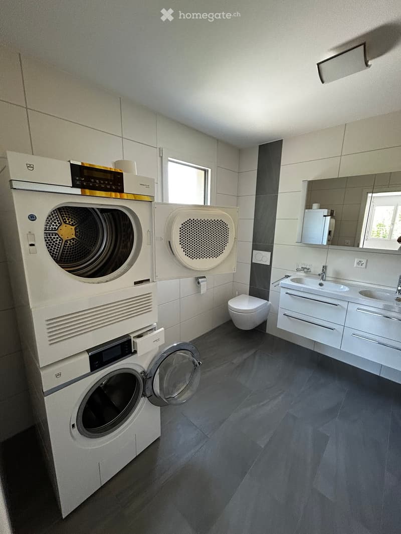 Modern bathroom with white tiled walls, a washing machine and dryer unit, a sink with a mirror, and a toilet.