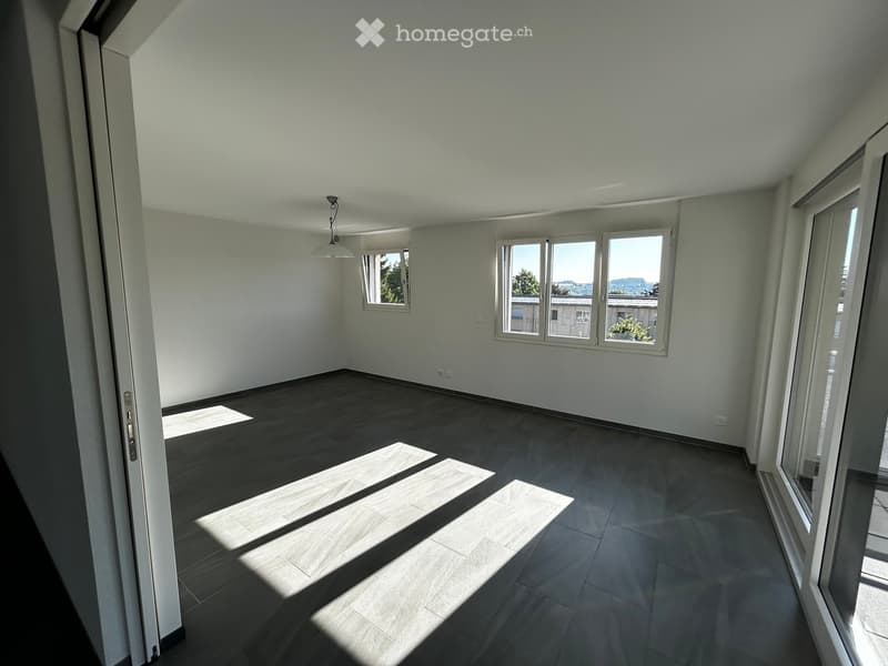 Empty room with wooden flooring, white walls, and large windows allowing natural light to enter. The space is unfurnished and appears to be a living or dining area.